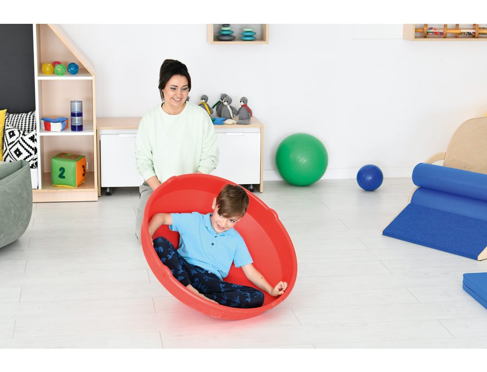 Dítě sedí v červené terapeutické misce během senzorické terapie, pod dohledem terapeuta.
