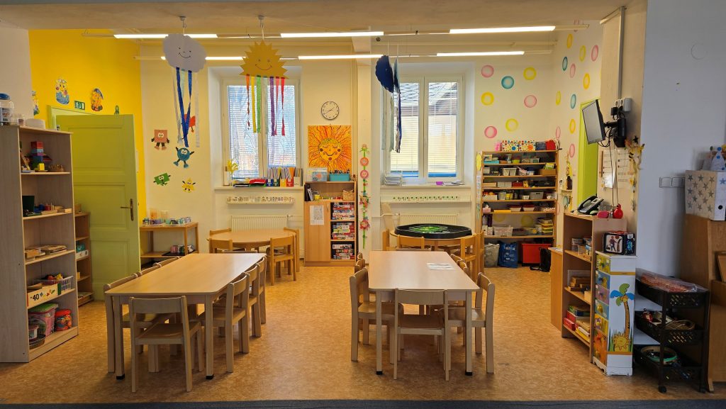 Barevně dekorovaná třída mateřské školy s dřevěným nábytkem, regály plnými výtvarných potřeb a zavěšenými papírovými dekoracemi.