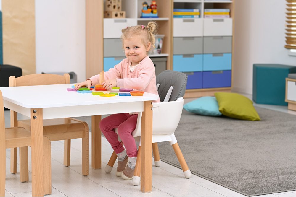 Na obrázku je malá holčička sedící u stolu, kde si hraje s barevnými kostkami. Má na sobě růžové tričko a kalhoty, usmívá se a vypadá velmi spokojeně. V pozadí je dětský pokoj s nábytkem v pastelových barvách.