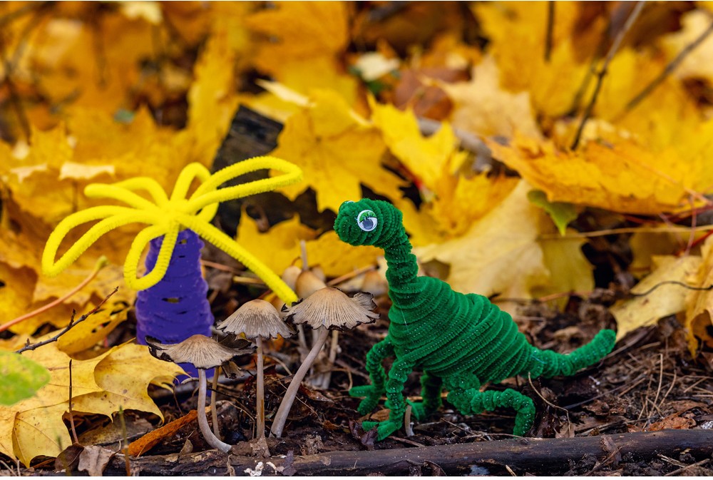 Dinosaurus z kreativních drátků, vyrobený z různých barevných materiálů, stojí na pozadí přírodního prostředí.