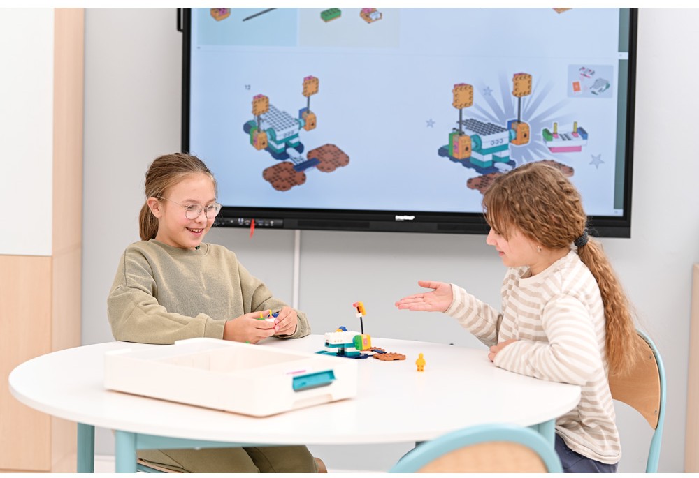 Dívky se soustředí na stavbu modelu z LEGO, zobrazeného na obrazovce v pozadí, a aktivně spolupracují při dokončení projektu.