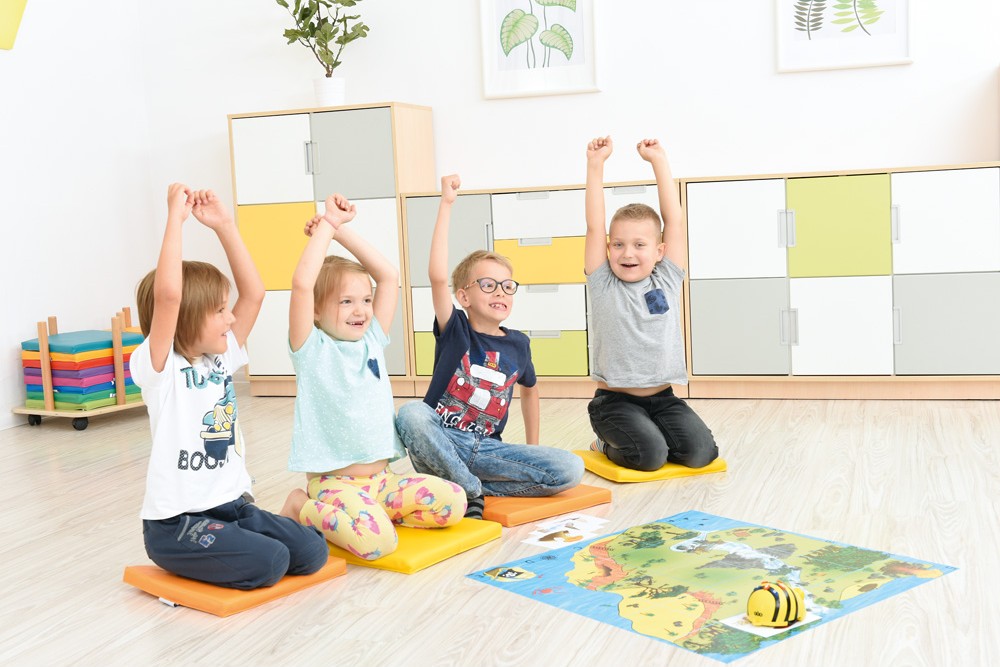 Děti sedí na podložkách ve třídě a hrají si s programovatelným robotem Bee-Bot na vzdělávací mapě.