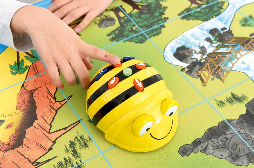 Detail interaktivního robota Bee-Bot na barevné vzdělávací podložce s dětskou rukou.