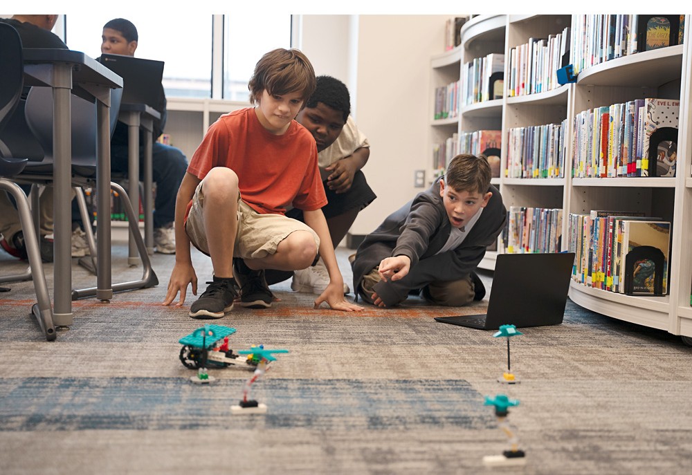 Děti v knihovně sledují a staví LEGO roboty na zemi, ukazují a diskutují o svých konstrukcích.