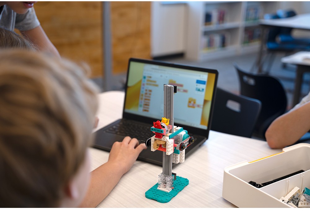 Učitelka dohlíží na stavění a programování LEGO modelů, všichni jsou zapojení a usměvaví.