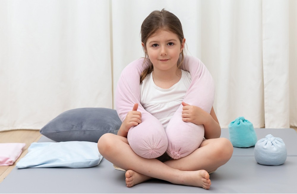 Dívka sedí na zemi, drží relaxační polštáře kolem krku, různé polštáře a pytlíky kolem ní