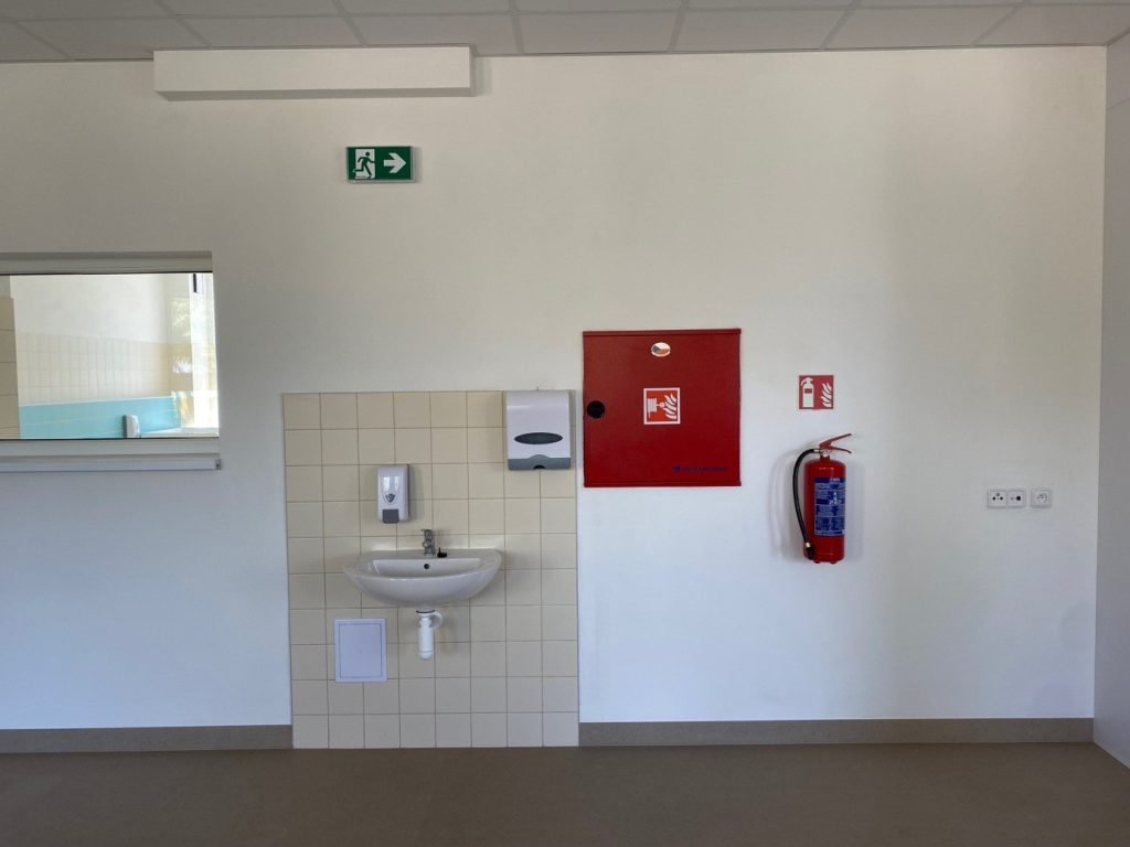 Pôvodný priestor v škôlke s umývadlom, výdajným okienkom, hasiacim prístrojom a voľnou stenou pred plánovanou úpravou interiéru.