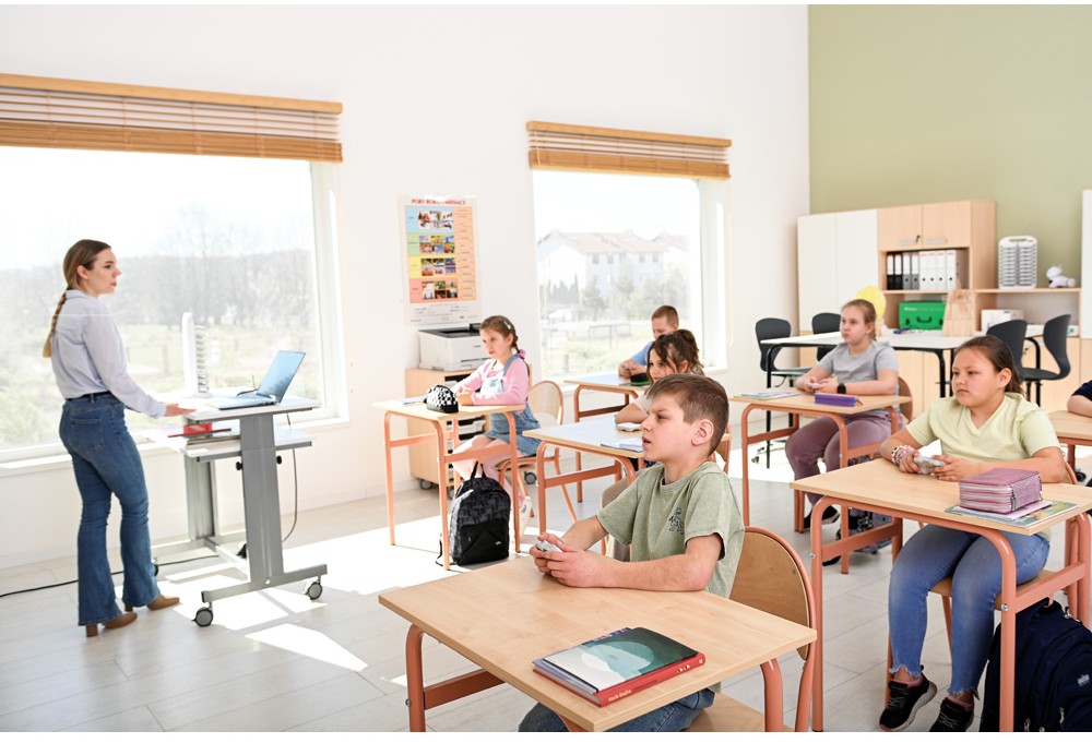 Učitelka vede vyučování ve světlé školní třídě vybavené moderními lavicemi a židlemi.